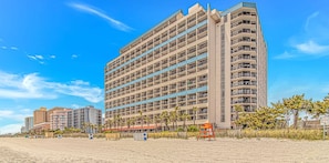 Un hotel de varios pisos con balcones, situado cerca de una playa con palmeras.