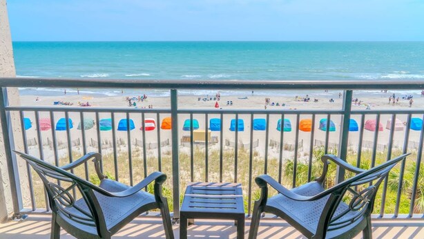 Un balcon avec deux chaises et une petite table, donnant sur une plage parsemée de parasols colorés et animée par la présence de nombreuses personnes.