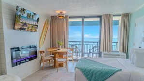 Une chambre d’hôtel comprenant un lit, une télévision, une table à manger avec des chaises, un balcon avec vue sur la mer et une planche de surf.