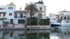 A marina with boats docked, a white building with a stone facade, and a palm tree in the foreground.