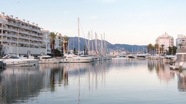 A marina with multiple yachts docked, a building with balconies, and a mountainous backdrop.