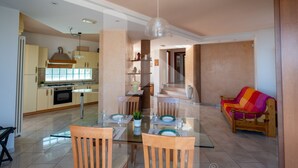A modern living room with a glass dining table, wooden chairs, a colorful sofa, and a kitchen in the background. - Homiday - Villa Moretti (Colonnella)