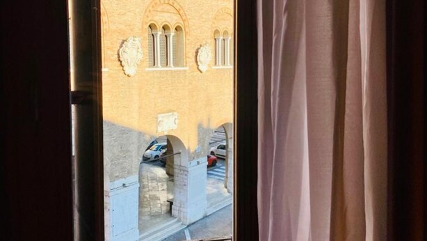 A view through a window with a curtain, featuring a building with arched windows and a stone archway. - SUITE300 - Apartment in the heart of Treviso (Treviso)