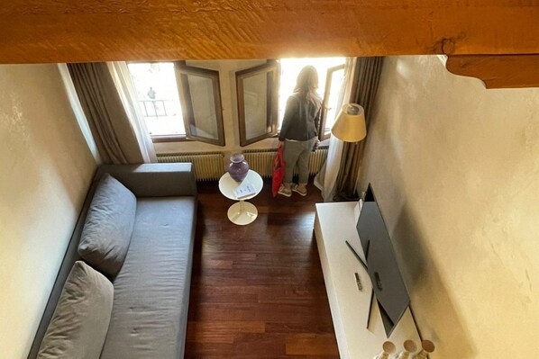 Ein kompakter Wohnbereich mit einer Couch, einem kleinen runden Tisch und einer Lampe. Ein Fenster mit Vorhängen bietet einen Ausblick.