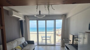 Ein großzügiger Wohnbereich mit Sofa, einem kleinen Tisch und einem TV-Schrank. Ein großes Schiebeglasfenster führt auf einen Balkon mit Meerblick.