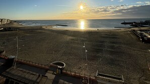 Blick auf einen Strand, bewölkter Himmel, eine untergehende Sonne über dem Meer und ein sandiger Strandabschnitt mit ein paar Gebäuden in der Ferne.