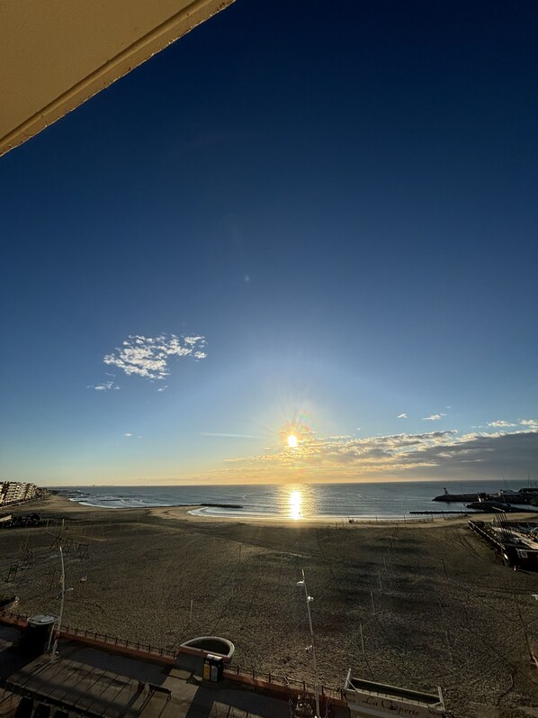 Strandudsigt med klar himmel, en solnedgang over havet og en sandstrand med få bygninger i det fjerne.