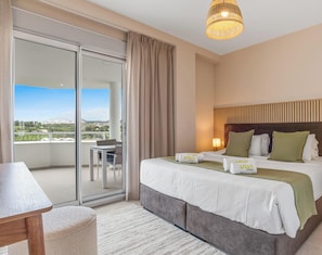 A hotel room with a bed, a table, a chair, a balcony with a view, and a hanging light fixture.