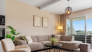 A modern living room with a sofa, a small table, and a potted plant. There is a balcony with a view of the city.