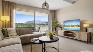 A modern living room with a sofa, a round coffee table, a TV, and a balcony with a view of the landscape.