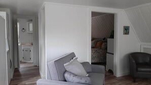 Una habitación con un sofá gris, un sillón oscuro y una cama con ropa de cama estampada. En la pared hay un cuadro pequeño.