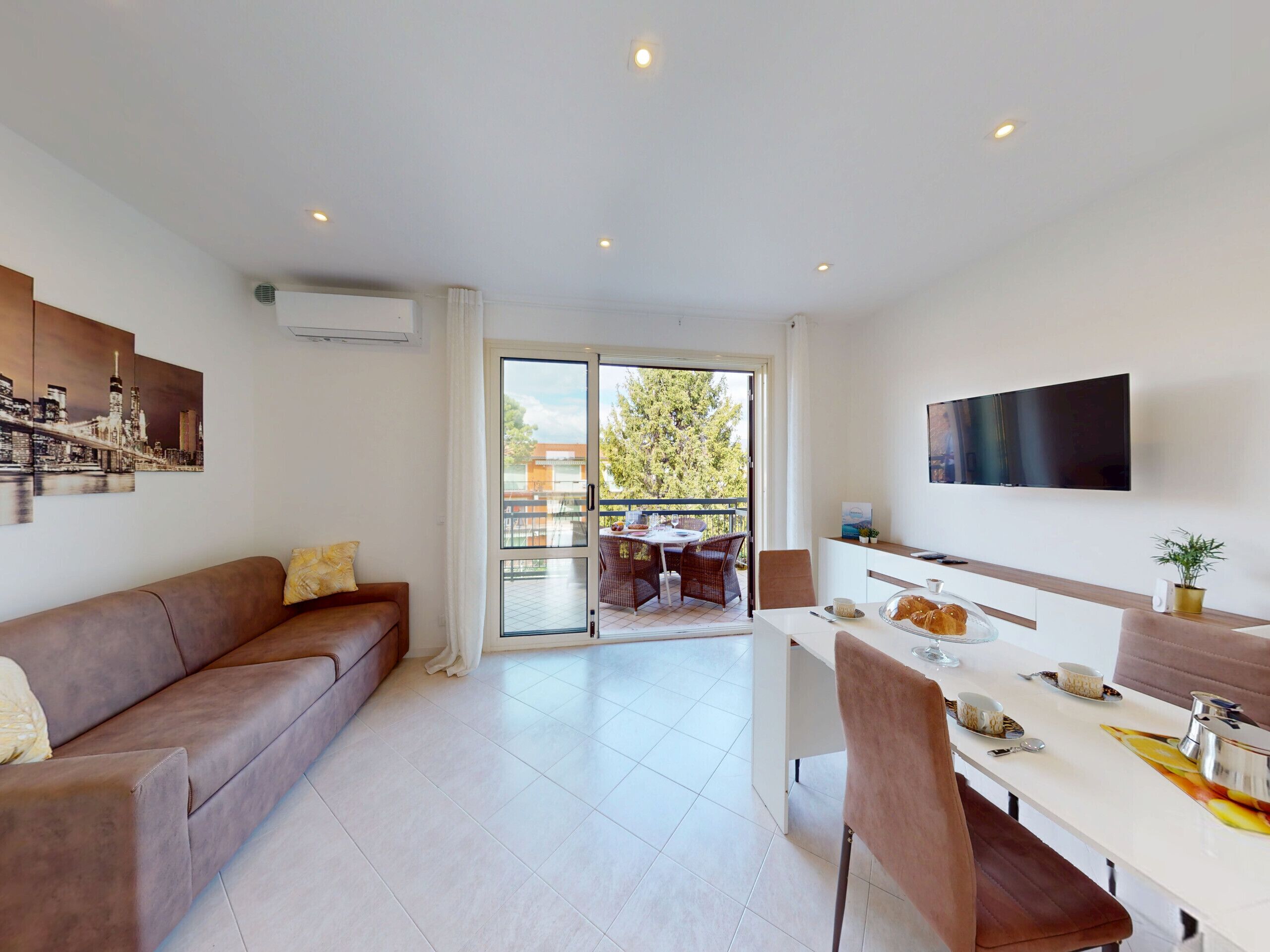 A modern living room with a sofa, a dining area with a table set for two, and a balcony with a view of trees and buildings.