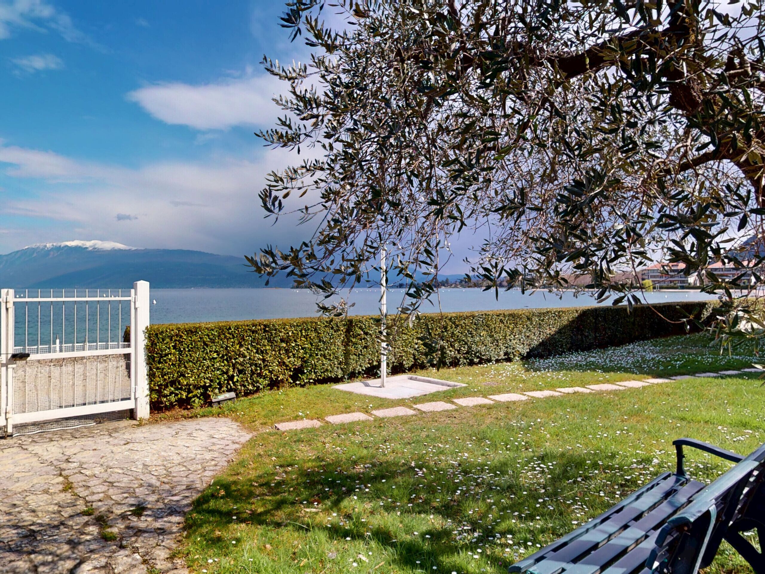 Ein Park mit einer Bank, einem Heckenstreifen und Blick auf einen See und Berge.