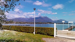 A scenic view of a lake with mountains in the background, a white fence, and a pathway leading to a dock. - Casa Marisol by Bookinggardalake (Manerba del Garda)