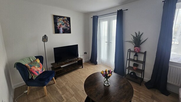 Un salón con un sillón azul oscuro, una mesa de centro de madera con un jarrón de flores, un televisor de pantalla plana sobre un mueble de madera y un ventanal con cortinas azules.