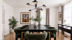 A dining area with a dark wooden table, green upholstered chairs, a white vase centerpiece, and a modern chandelier.