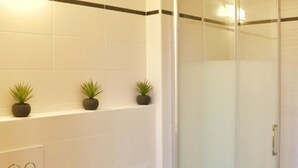 Un baño con azulejos blancos, tres plantas en macetas sobre una repisa, un box de ducha de cristal y una puerta con pomo.