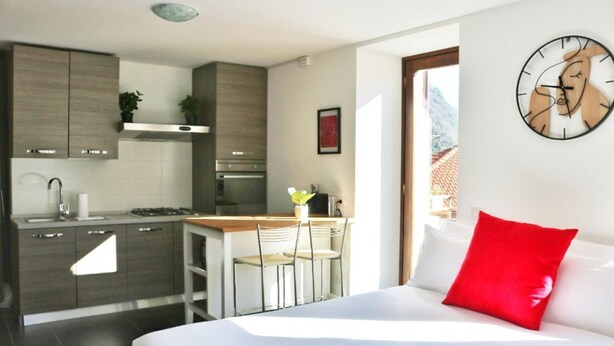 Una habitación de hotel moderna con zona de cocina, una cama con ropa de cama blanca y un almohadón rojo, y un reloj de pared.