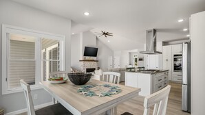 A modern kitchen with white cabinetry, a dining table with chairs, a TV mounted above the fireplace, and a ceiling fan. - The Lighthouse | Luxury Waterfront Retreat w/ Dock, Fireplace, Pools, Marina, & Golf (Greenbackville)