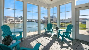 A sunlit room with large windows overlooking a body of water, featuring several turquoise Adirondack chairs and a ceiling fan. - The Lighthouse | Luxury Waterfront Retreat w/ Dock, Fireplace, Pools, Marina, & Golf (Greenbackville)
