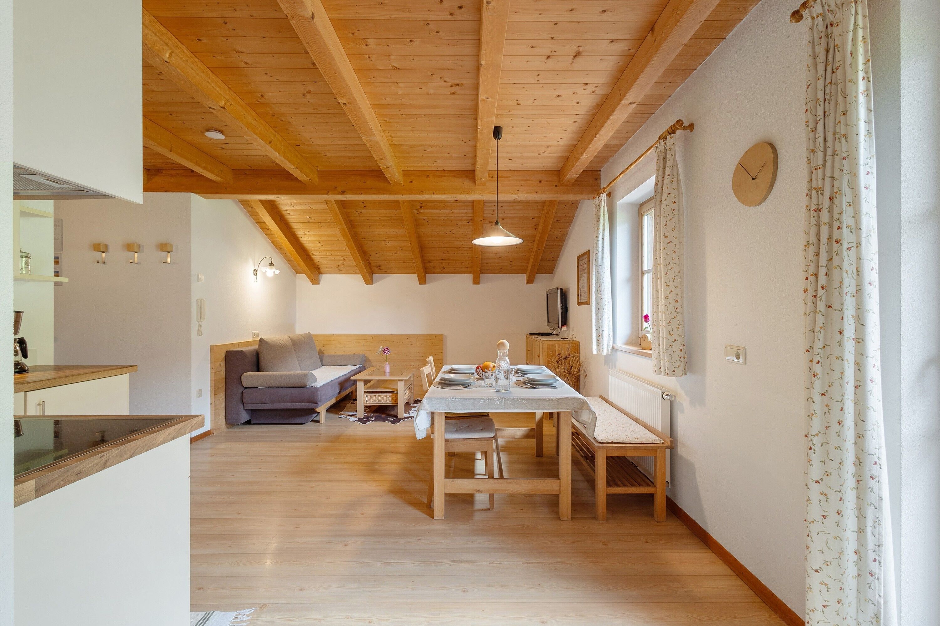 A room with a wooden ceiling, a dining table with chairs, a sofa, and a window with curtains.
