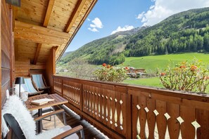Un balcon en bois offrant une vue sur un paysage montagneux, une table avec des chaises et un banc blanc et moelleux.