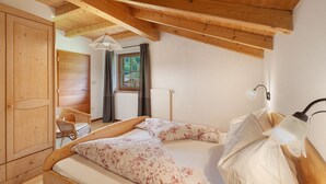 Sostre de fusta, llit amb roba de llit floral, una cadira i una finestra amb cortines.