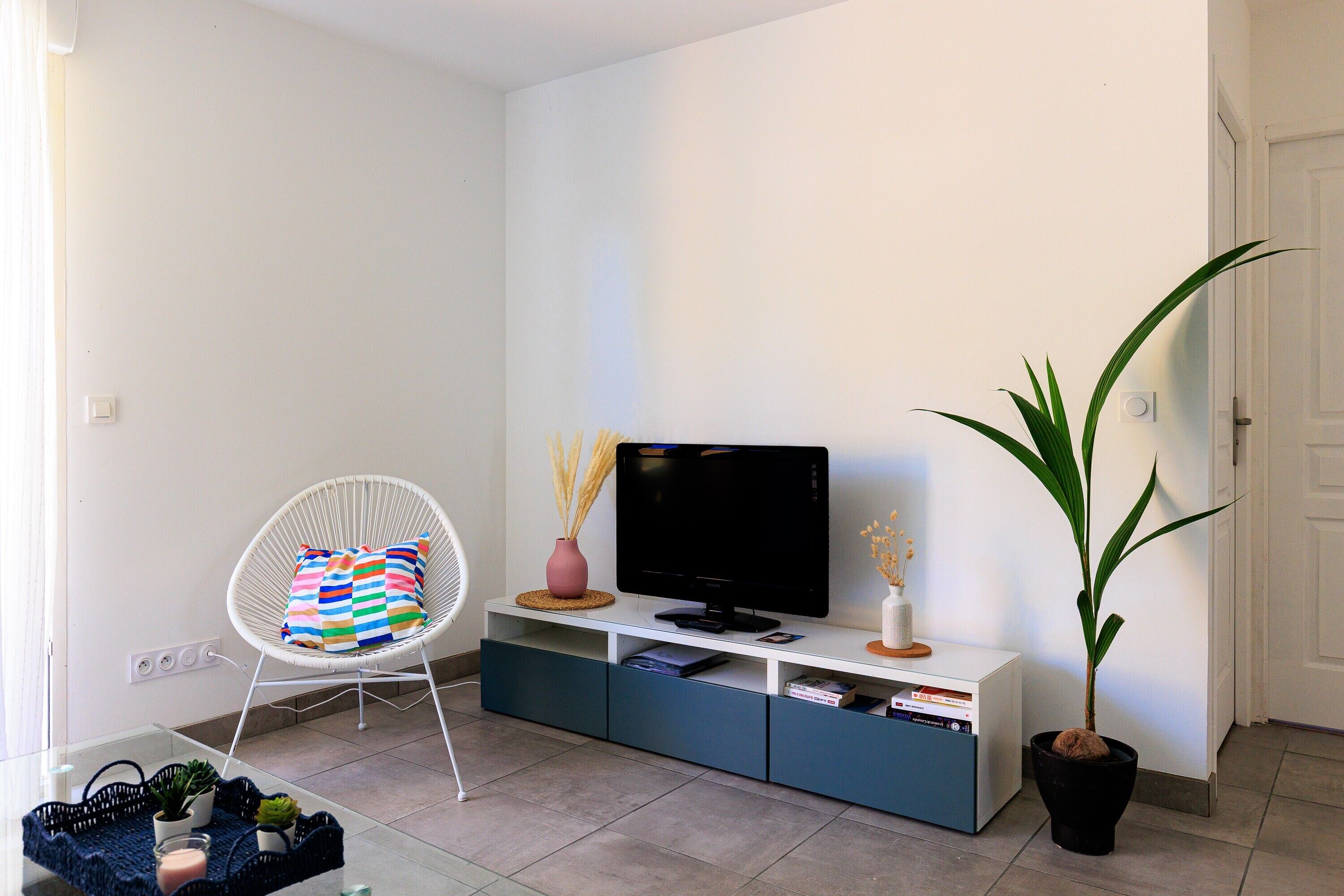Un salon avec un fauteuil blanc, un téléviseur posé sur un meuble, une plante en pot et une table basse en verre.