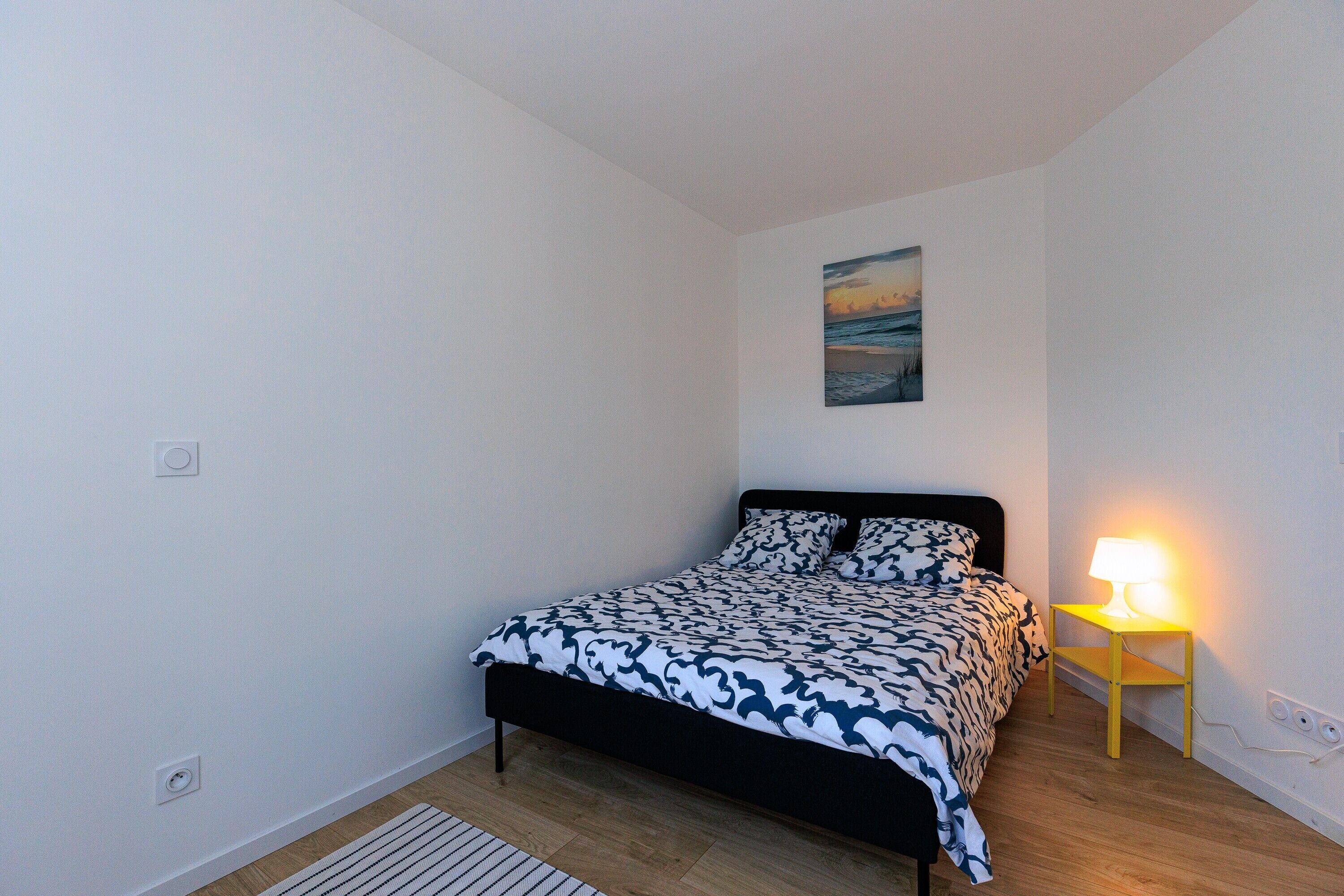 A bedroom with a bed, a bedside table with a lamp, a painting on the wall, and a radiator.