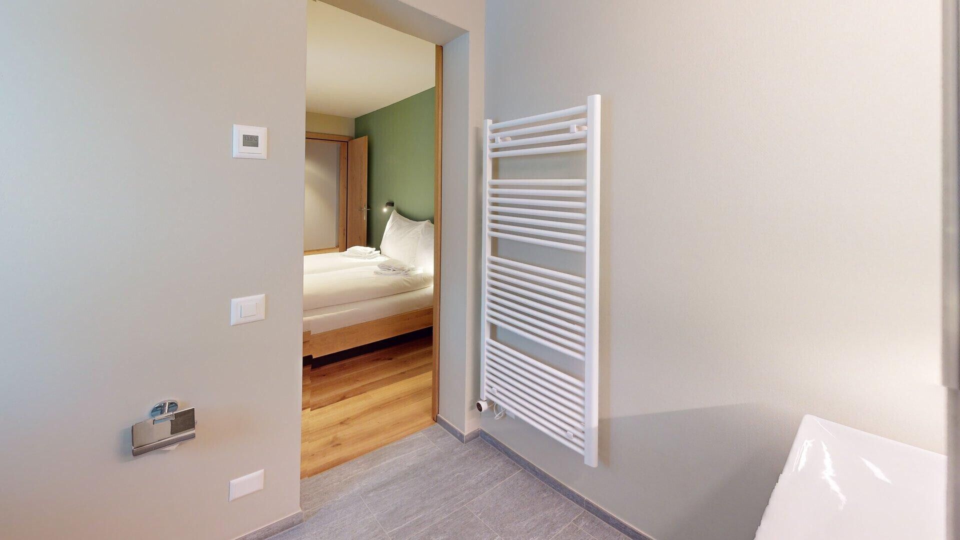 A hotel room with a wooden floor, a bed with white bedding, a white radiator, and a door leading to another room.