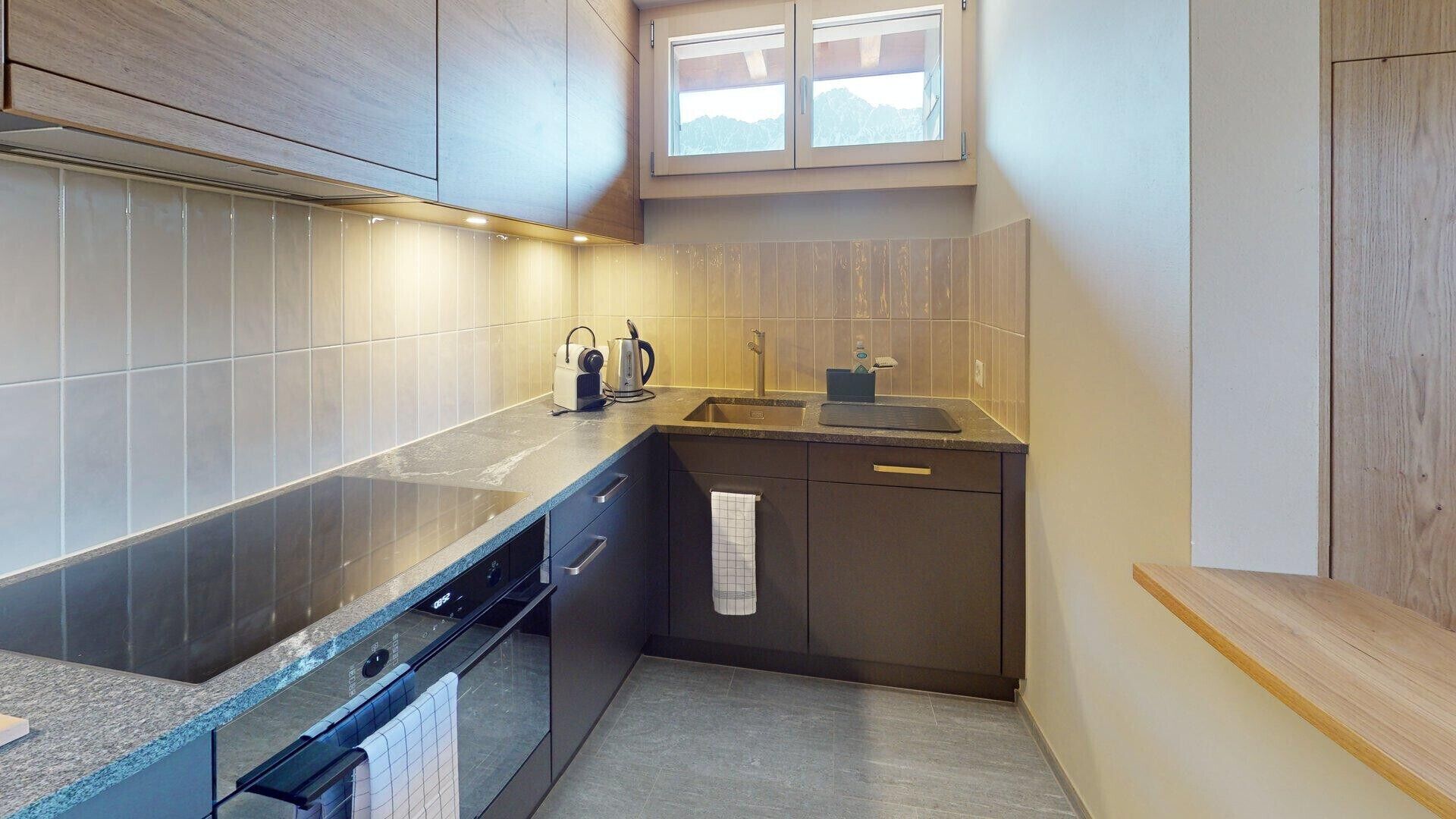 Eine kleine Küche mit dunklen Schränken, einem eingebauten Backofen, einem Spülbecken und einem Fenster über der Arbeitsfläche.