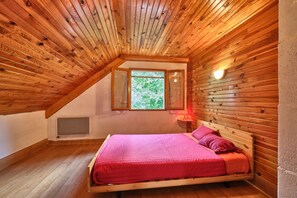 Et soveværelse med skråvæg, trævægge, en seng med pink sengetøj, et vindue og en lampe monteret på væggen.