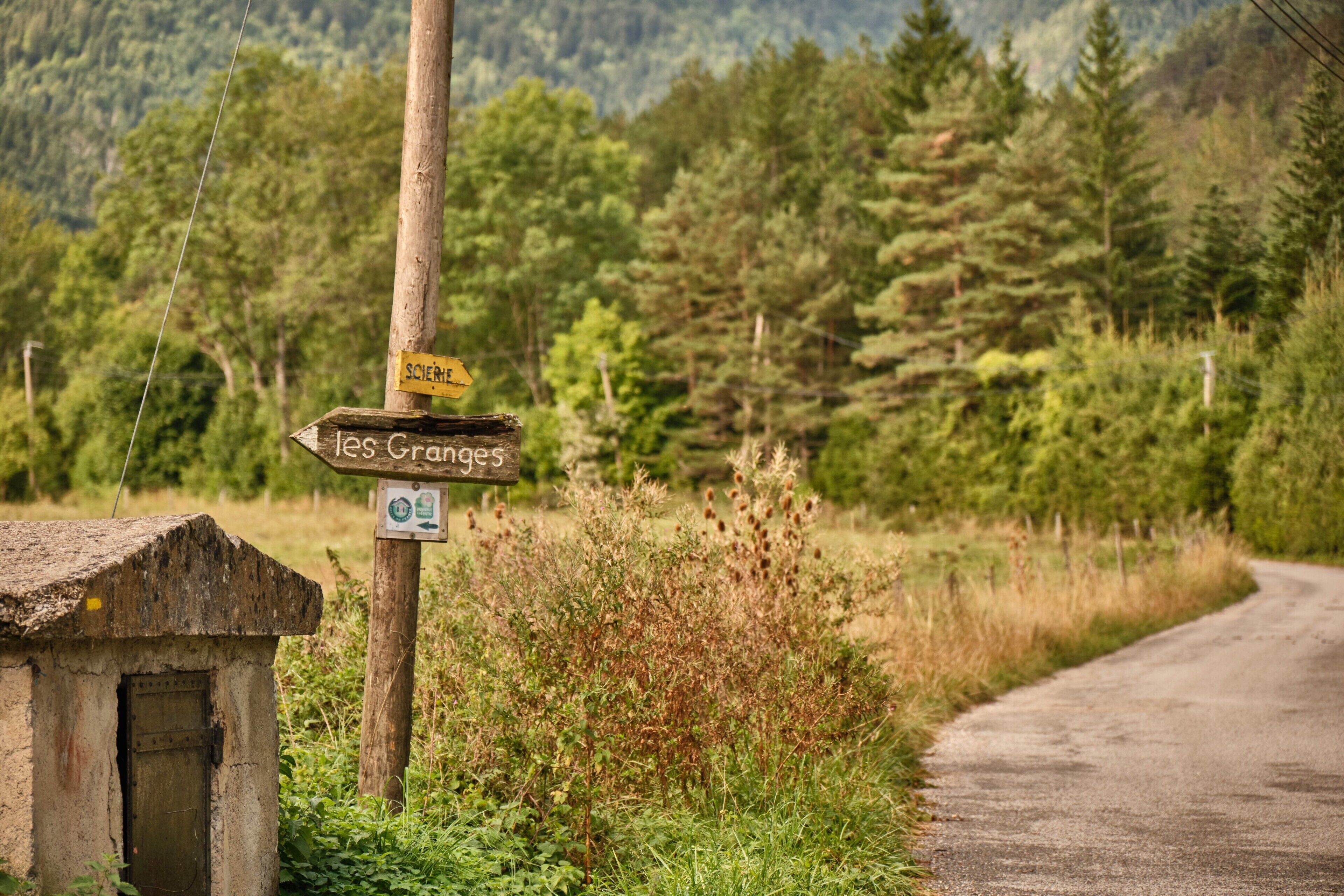 A rural road with a wooden signpost indicating directions to 'Les Granges' and 'SCIERIE'.