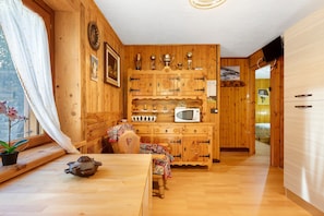 A wooden interior with a kitchen area, a dining table with a chair, and a window with curtains.