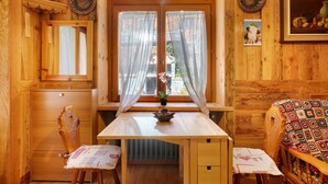 Interior de una cabaña de madera con una mesa de madera, dos sillas, una ventana con cortinas traslúcidas y un cuadro en la pared.