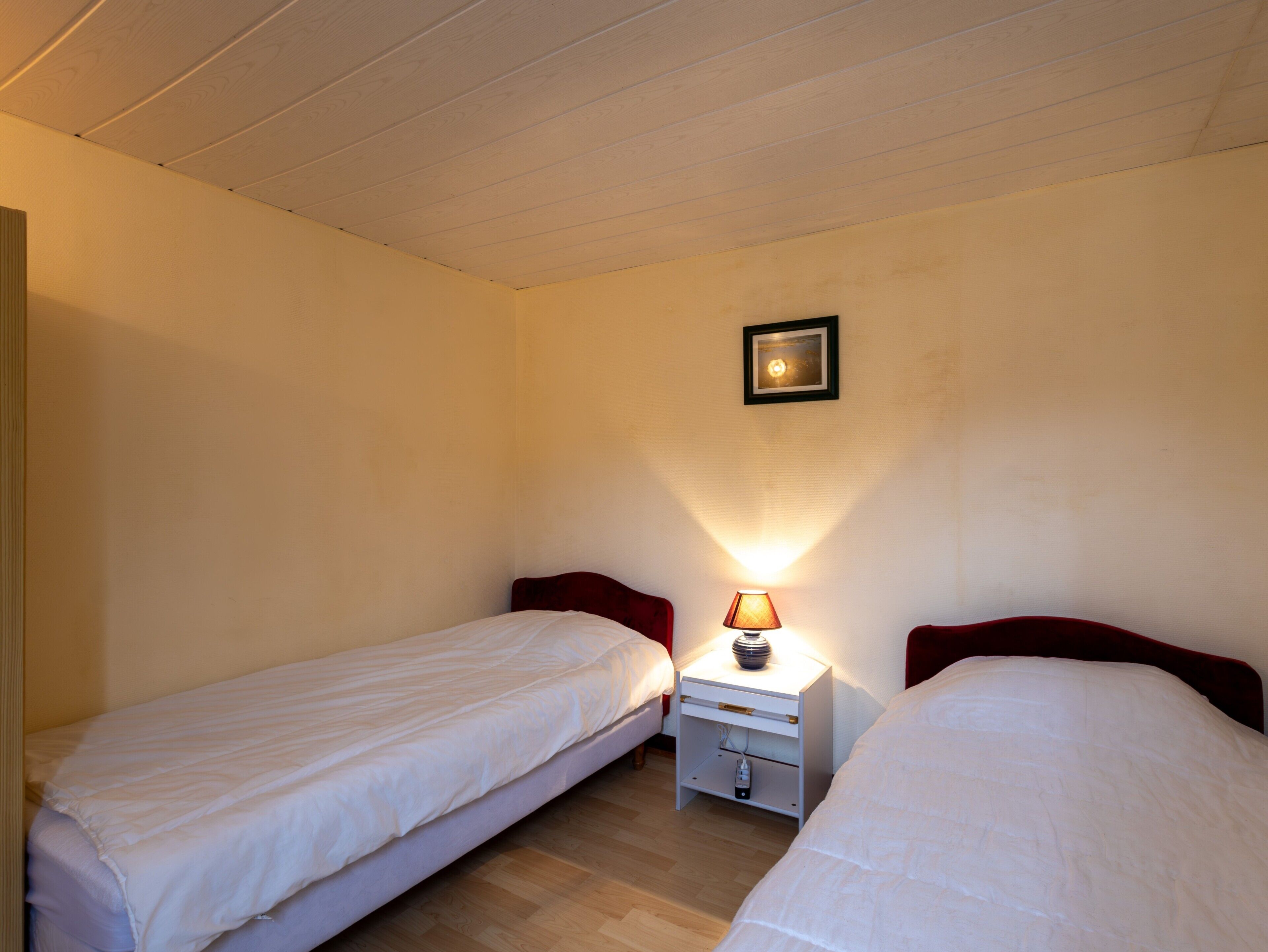 A small, single-room hotel room with two single beds, a nightstand with a lamp, and a framed picture on the wall.