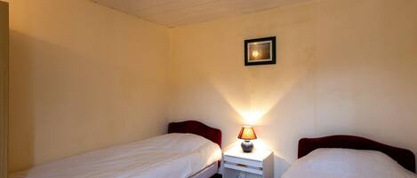 A small, single-room hotel room with two single beds, a nightstand with a lamp, and a framed picture on the wall.