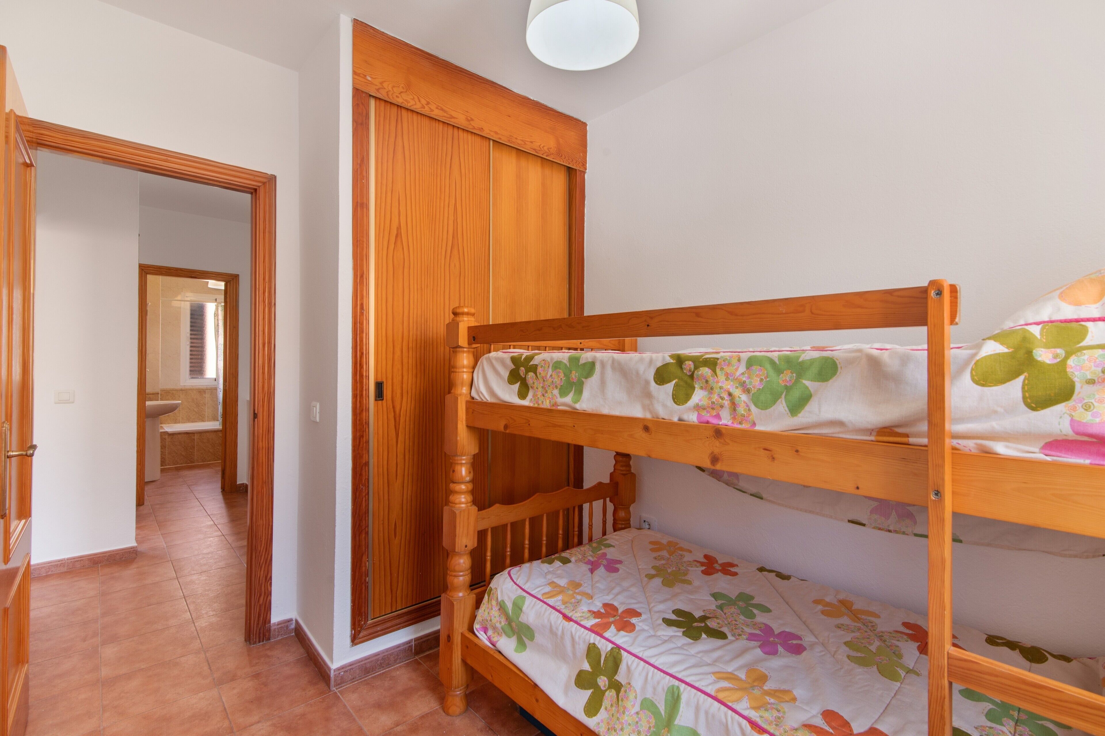 Un lit superposé avec une literie à motifs floraux, une armoire en bois et une salle de bain visible au fond.