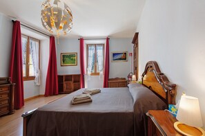 Ein Hotelzimmer mit einem großen Bett, einem hölzernen Kopfteil, einem Nachttisch aus Holz, einem Schreibtisch, einem Stuhl, einer Lampe und zwei mit Vorhängen versehenen Fenstern.