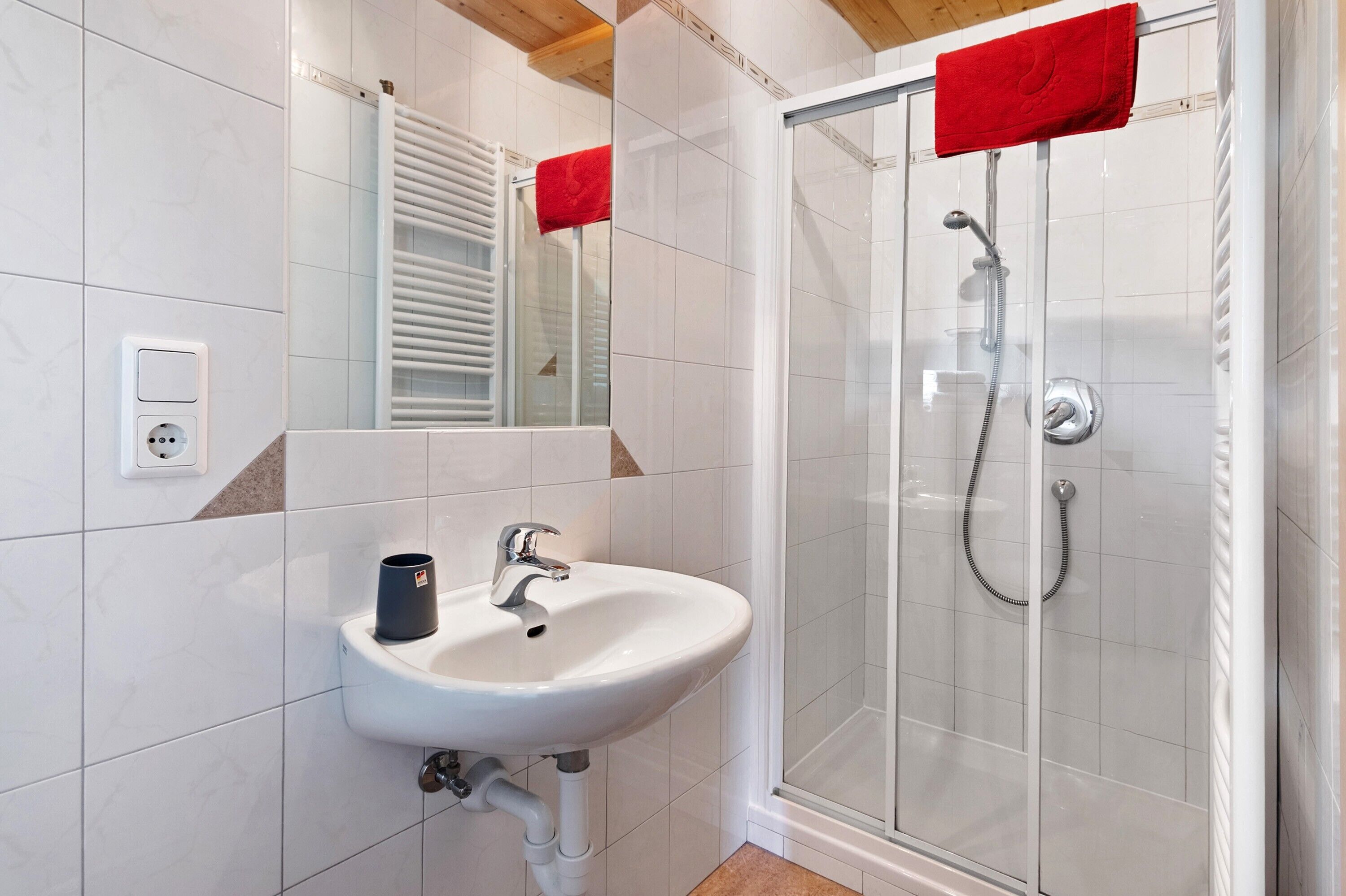Een badkamer met een witte wastafel, een spiegel, een douche met glazen deur en een rode handdoek aan de deur.