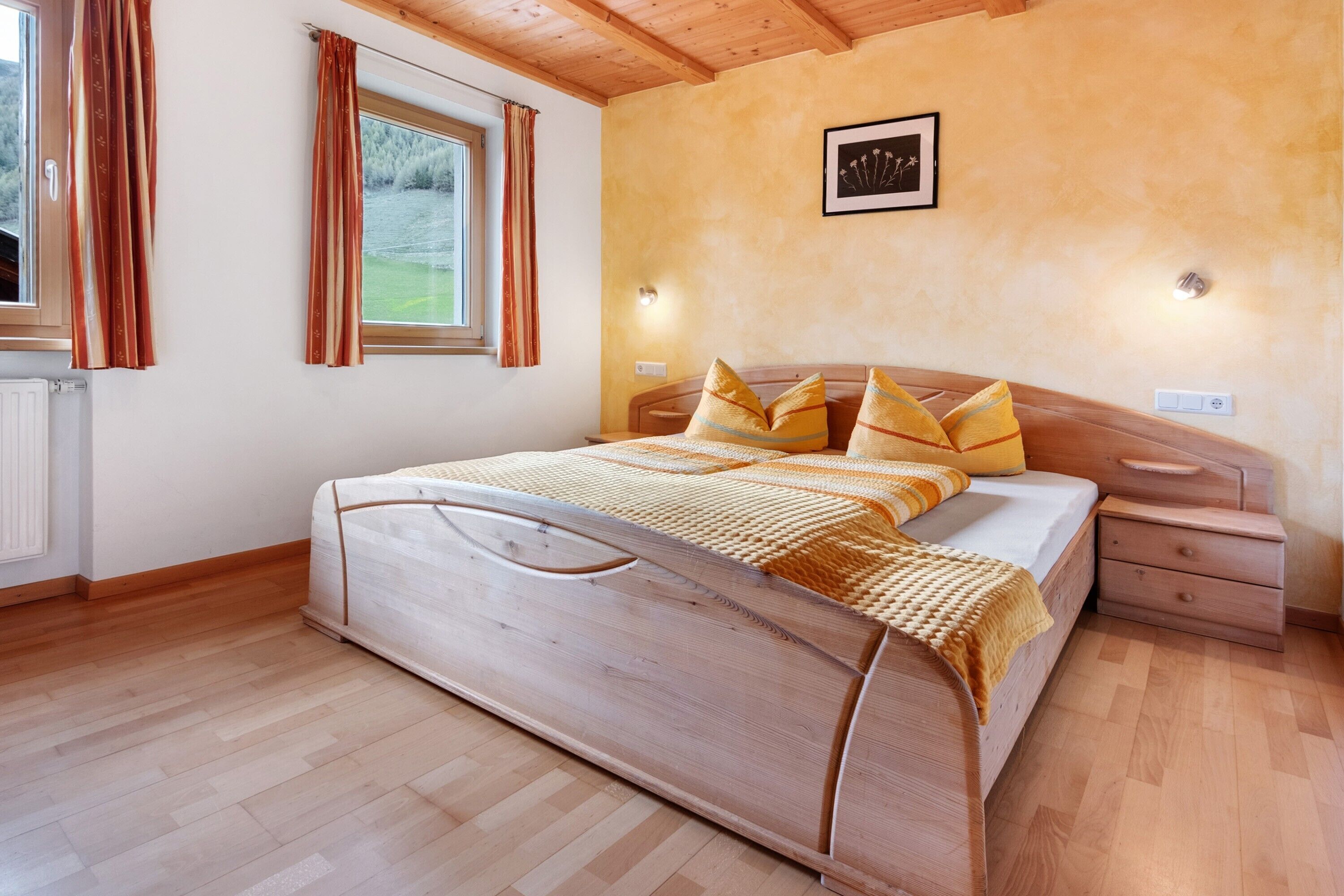 A wooden bed with a headboard, a bedside table, a nightstand, a window with curtains, and a wall-mounted light fixture.