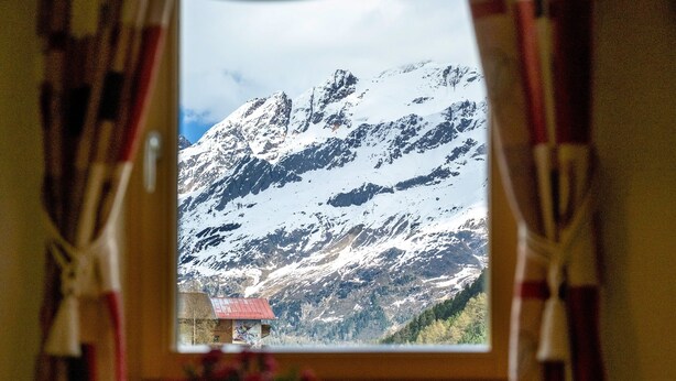 Vista de la montaña a través de una ventana con cortinas, una mesa con un vaso y un jarrón con flores.