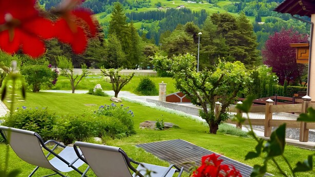 Une vue d’un jardin luxuriant avec une chaise et une table, entouré de verdure et avec une chaîne de montagnes qui se profile à l’horizon.