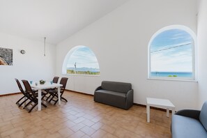A spacious room with a dining table, chairs, a sofa, and two arched windows offering a view of the sea.