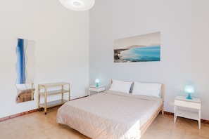 A bedroom with a bed, a bedside table, a nightstand, a wall-mounted shelf, and a painting of a coastal scene.