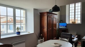 Un espace repas comprenant une table, des chaises, un meuble, un téléviseur et une fenêtre donnant sur des bâtiments.