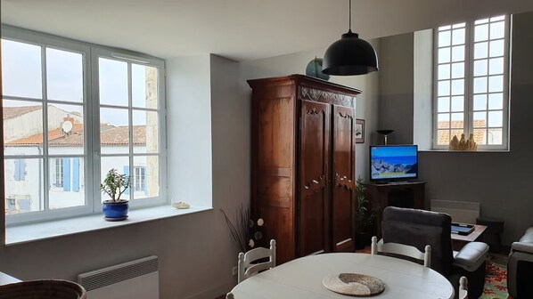 Un comedor con una mesa, sillas, un armario, un televisor y un ventanal con vistas a edificios.