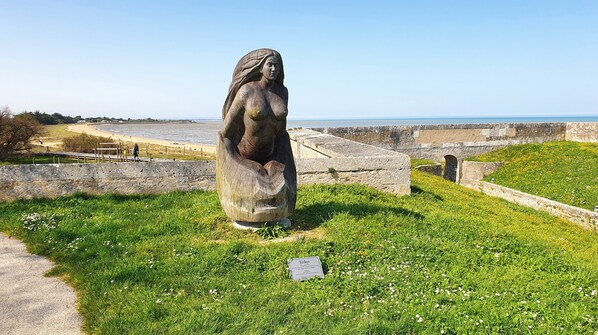 Una estatua de una mujer en un área con césped, una pared de piedra y vista despejada al mar.