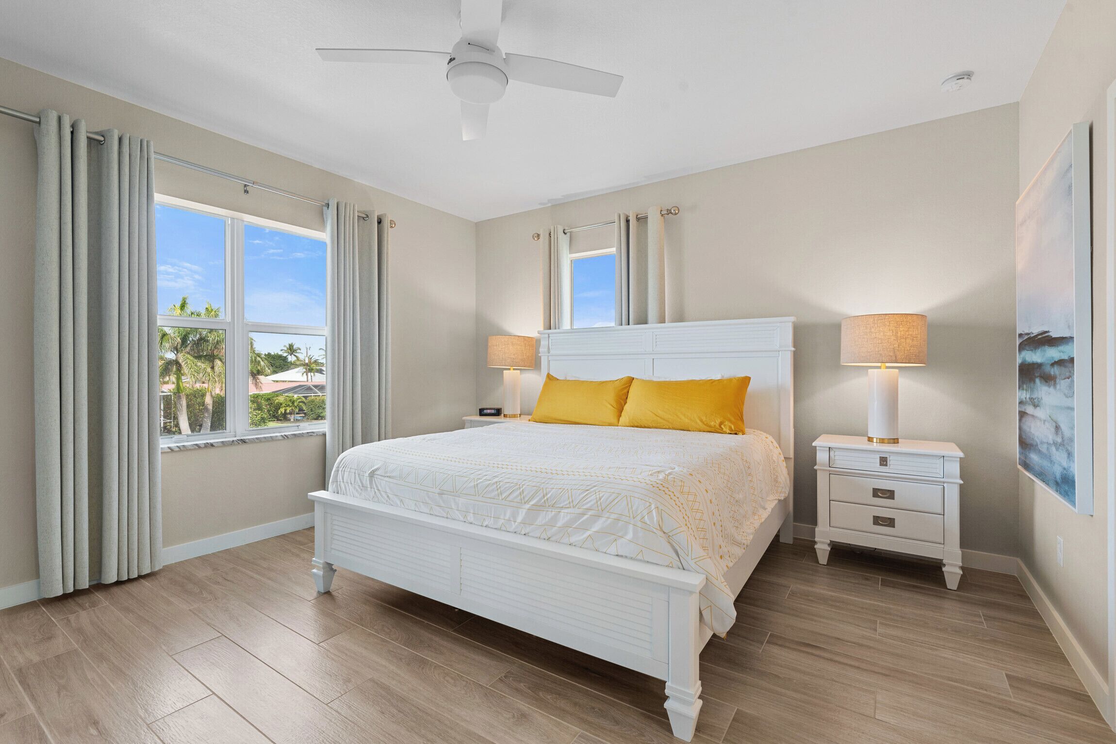 A bedroom with a white bed, two bedside tables with lamps, a ceiling fan, and large windows with curtains.