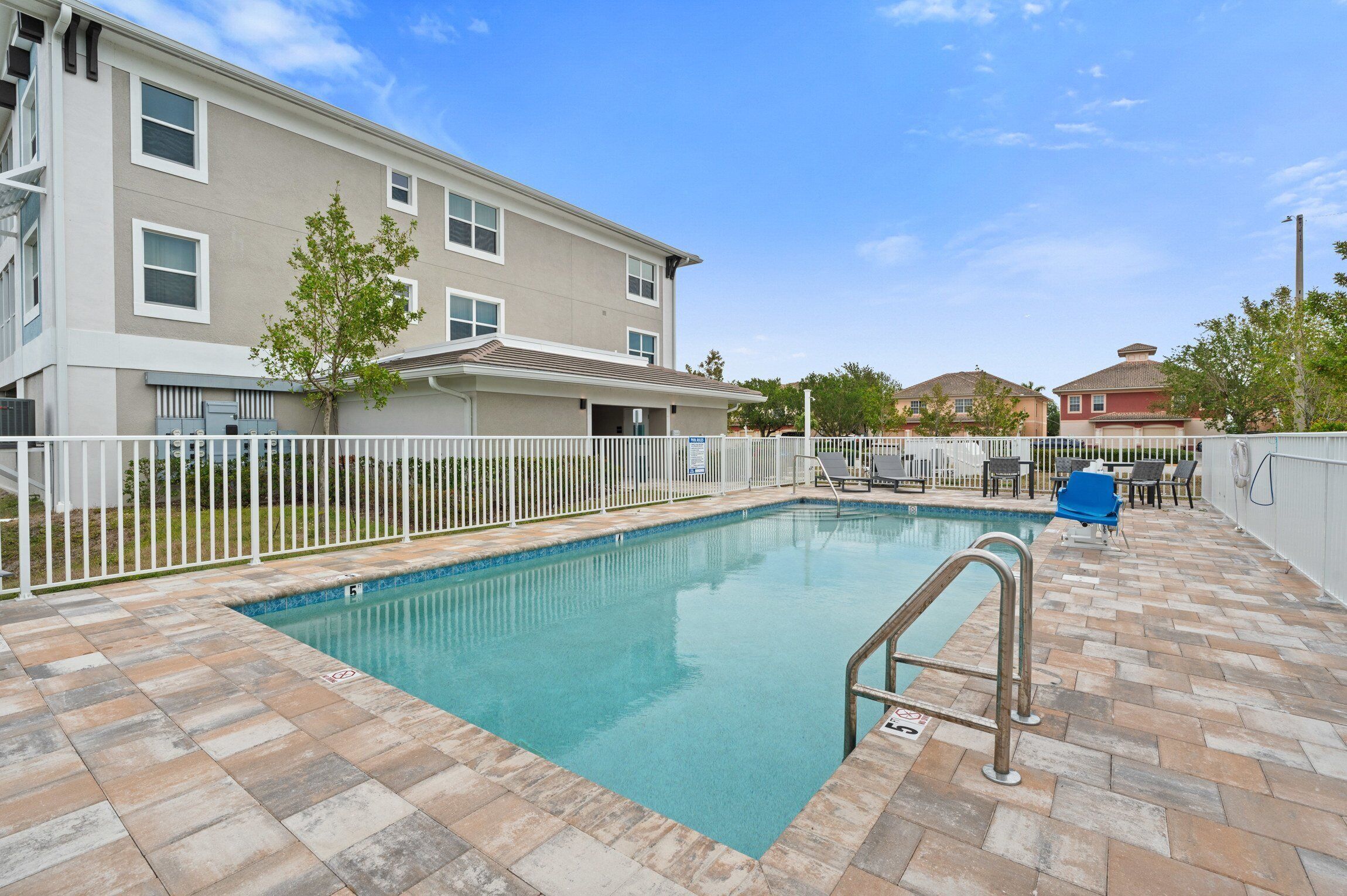A swimming pool with a tiled deck, a white fence, and a building with multiple windows in the background.